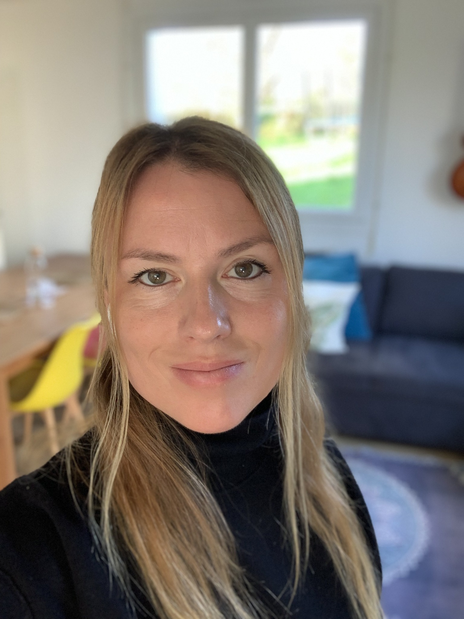 A woman with long blonde hair wearing a black turtleneck, smiling and posing for a selfie in a well-lit room with a blurred background featuring a couch and a dining table.