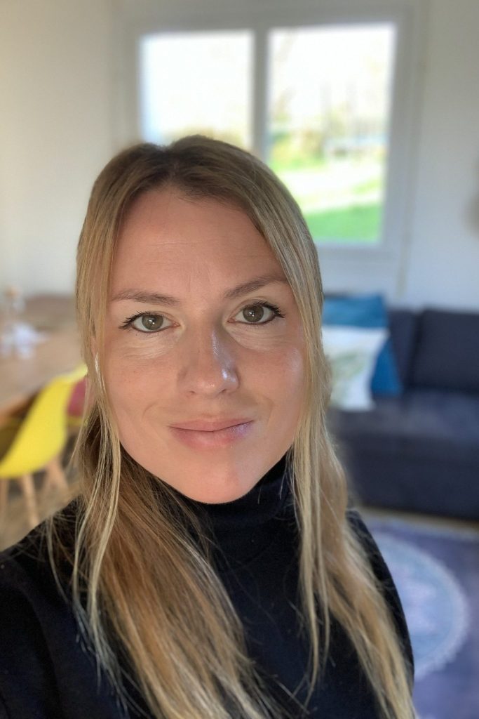 A woman with long blonde hair wearing a black turtleneck, smiling and posing for a selfie in a well-lit room with a blurred background featuring a couch and a dining table.